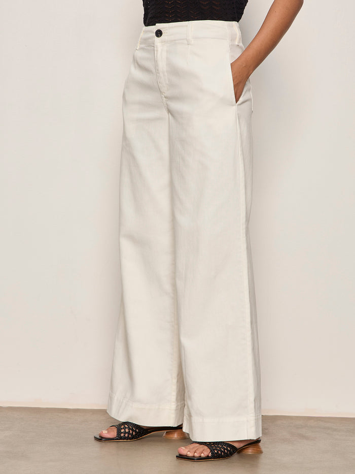 A person wearing Sanctuary Clothing’s academy wide leg collagen pants, styled with a black top and black open-toe sandals, stands against a plain light-colored wall.
