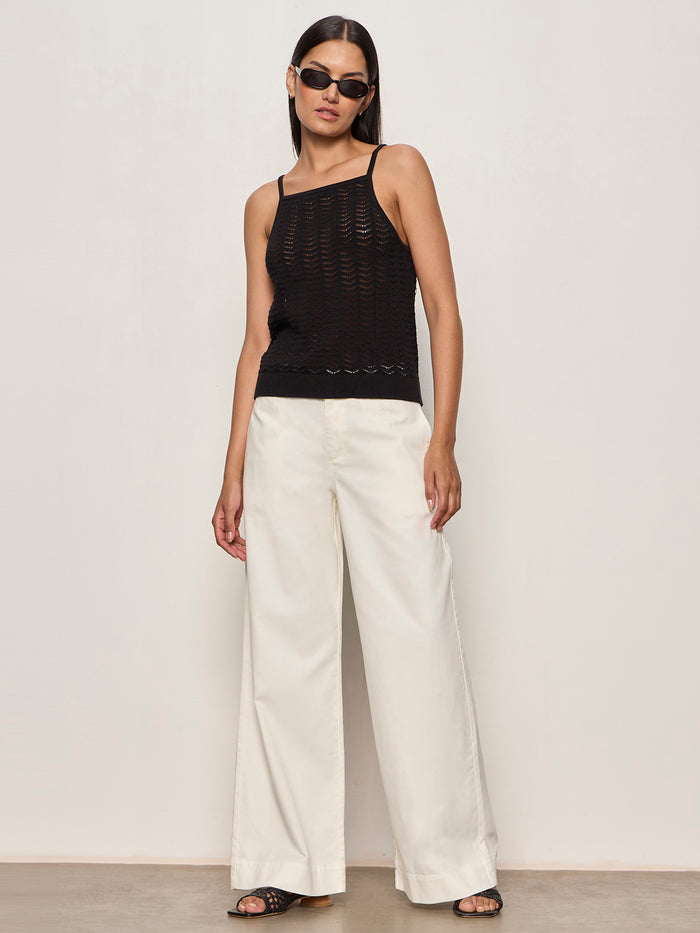 A woman wears Sanctuary Clothing's academy wide leg collagen pants, a sleeveless black knit top, black sandals, and sunglasses while standing against a plain light-colored wall.