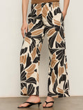 A person wears Sanctuary Clothing’s academy wide leg lotus trousers, featuring a bold black, beige, and brown abstract floral print, paired with black open-toe sandals. Their upper body is partially out of frame.