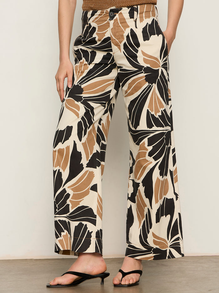 A person wears Sanctuary Clothing’s academy wide leg lotus trousers, featuring a bold black, beige, and brown abstract floral print, paired with black open-toe sandals. Their upper body is partially out of frame.