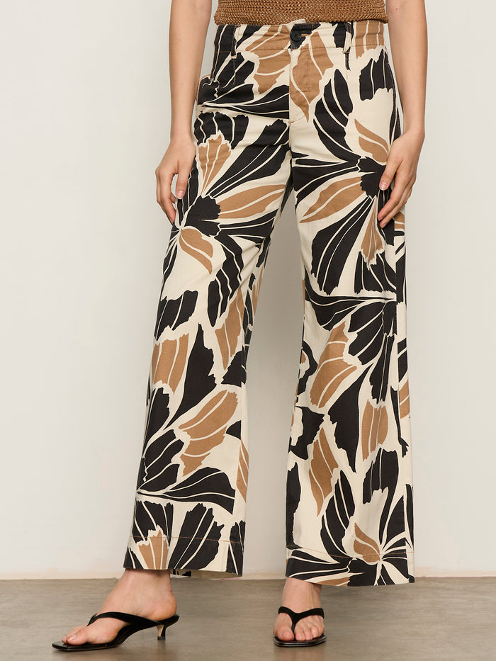 A woman models the Sanctuary Clothing "academy wide leg lotus" pants, featuring a high-waisted, wide-leg silhouette and bold abstract leaf print in black, brown, and beige tones, paired with black open-toe heeled sandals.