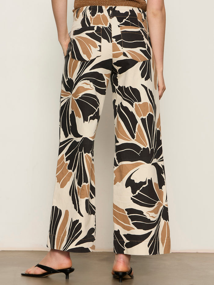 A person stands facing away, wearing Sanctuary Clothing's academy wide leg lotus pants featuring large black and brown abstract floral prints and black heeled sandals, against a plain light background.