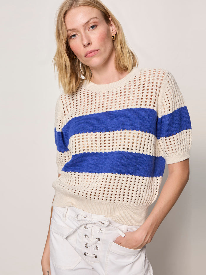 A woman with blonde hair wears the Sanctuary Clothing beachside openwork tee collagen w luminous blue, a cream short-sleeve crochet sweater with bold blue stripes, paired with high-waisted white lace-up front pants against a plain light background.