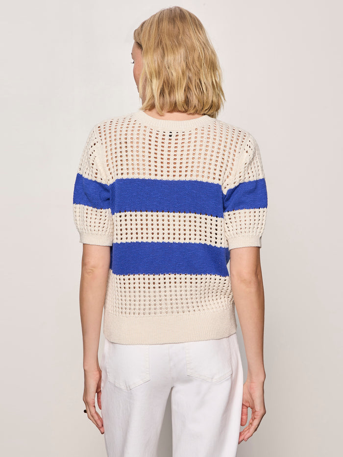 A person with blonde shoulder-length hair is shown from the back, wearing Sanctuary Clothing's beachside openwork tee collagen w luminous blue and white pants, standing against a plain light background.