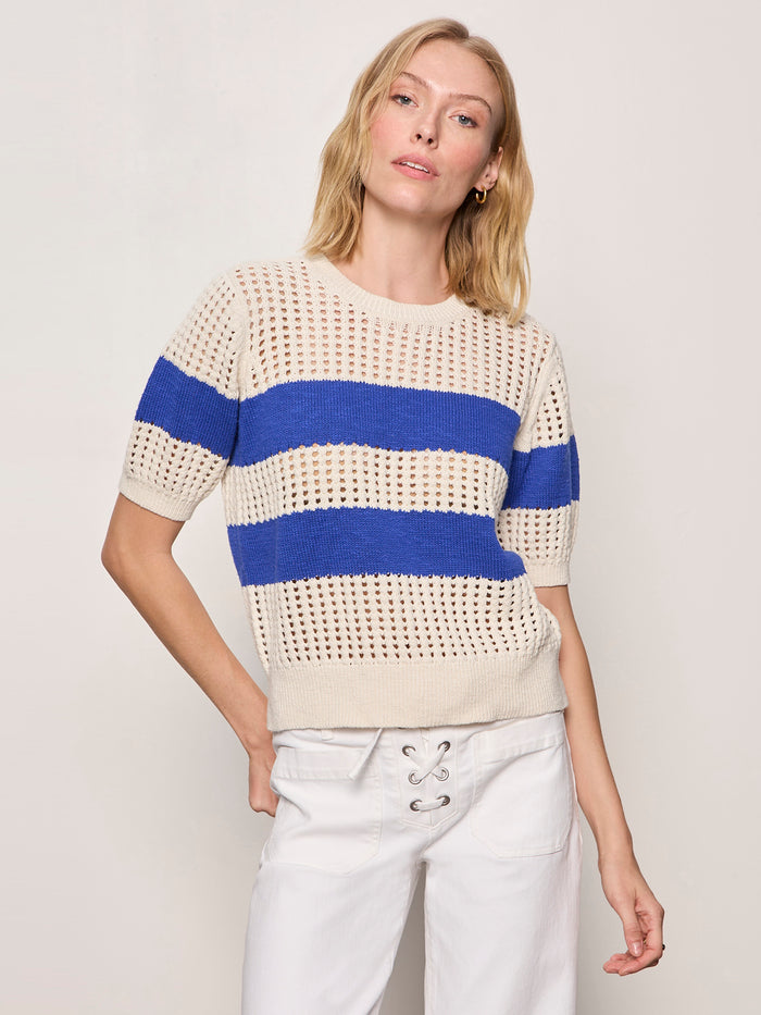 A woman with blonde hair wears the Sanctuary Clothing beachside openwork tee collagen w luminous blue— a short-sleeved, cream knit sweater with two wide blue stripes—paired with white lace-up waist pants against a plain light background.
