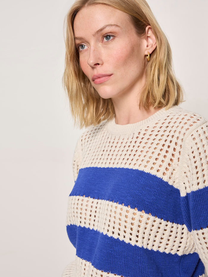 A woman with blonde hair and gold hoop earrings models the Sanctuary Clothing beachside openwork tee collagen w luminous blue, a beige open-knit sweater with bold blue stripes, as she gazes to the side against a plain background.