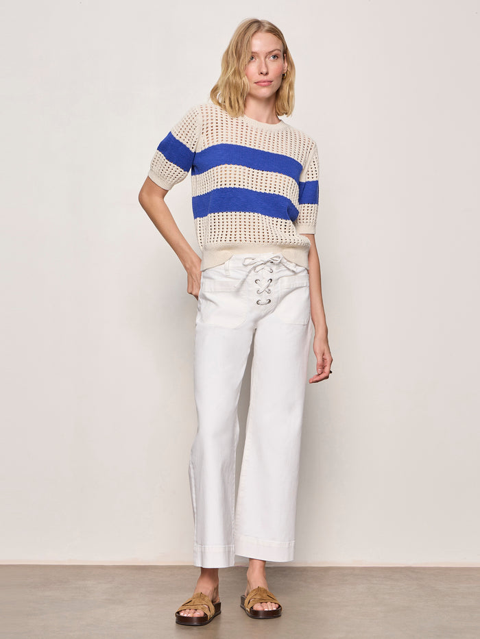 A woman with short blonde hair stands in a relaxed pose against a plain background, wearing the Sanctuary Clothing beachside openwork tee collagen w luminous blue and white wide-leg lace-up pants, paired with gold sandals.