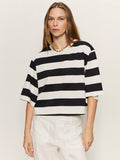 A woman with straight, light brown hair wears the Sanctuary Clothing belle tee in chalk and black stripes, paired with white pants and a gold chain necklace, standing against a plain light background.
