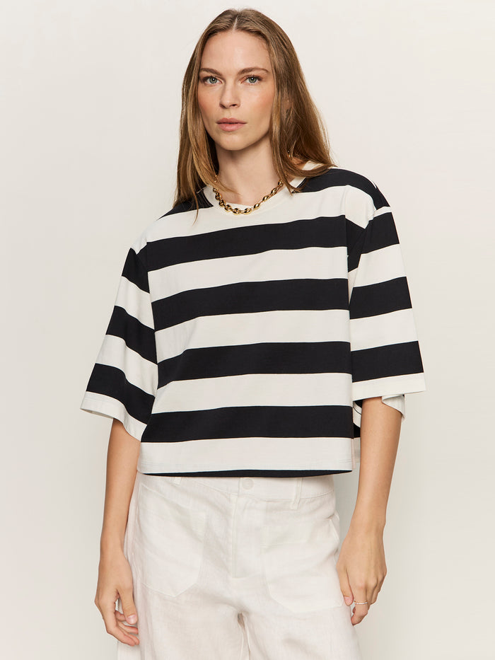 A woman with straight, light brown hair wears the Sanctuary Clothing belle tee in chalk and black stripes, paired with white pants and a gold chain necklace, standing against a plain light background.