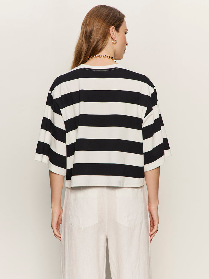 A person with long brown hair is seen from behind wearing the Sanctuary Clothing belle tee in chalk and black stripes, paired with white pants, against a plain light background.