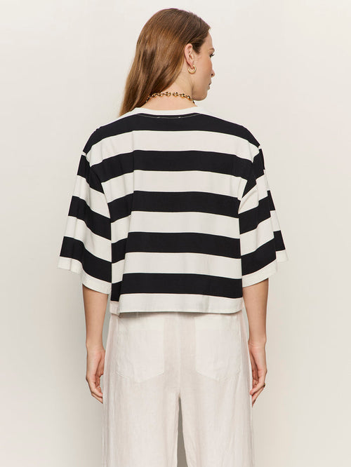 A person with long brown hair is seen from behind wearing the Sanctuary Clothing belle tee in chalk and black stripes, paired with white pants, against a plain light background.