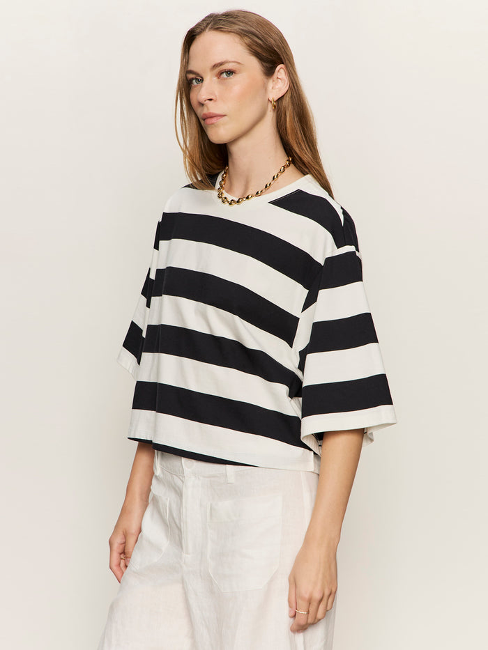A woman with long brown hair wears the Sanctuary Clothing belle tee chalk and black, white pants, and a chunky gold necklace, standing against a plain light background.