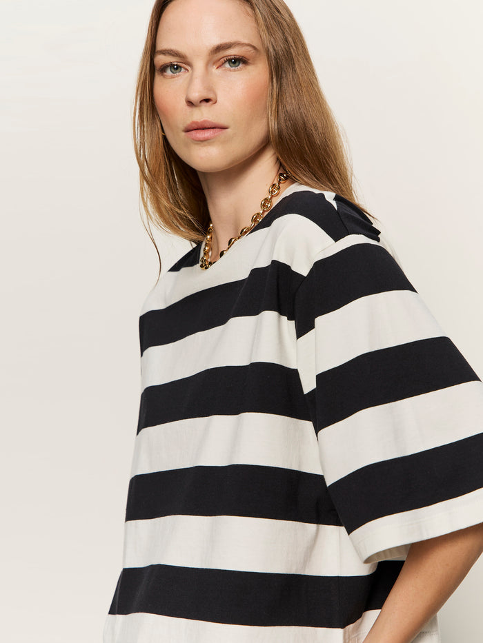 A woman with long light brown hair wears a chunky gold chain and the Sanctuary Clothing belle tee in chalk and black, featuring loose-fitting black and white horizontal stripes against a plain light background.