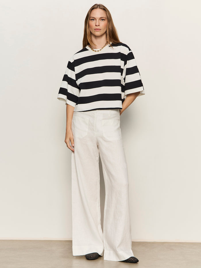 A woman with straight, long hair and a relaxed posture wears the Sanctuary Clothing belle tee in chalk and black, styled with wide-legged white pants, a gold necklace, and black shoes against a plain light background.