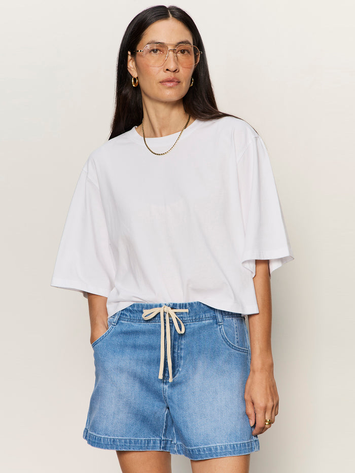 Woman with long dark hair wears clear glasses, a gold necklace, and blue drawstring denim shorts. She stands against a plain light background in the Sanctuary Clothing belle tee white.