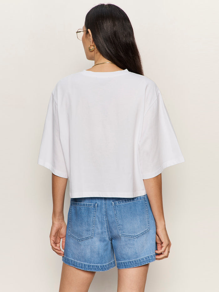 A woman with long dark hair and glasses stands with her back to the camera against a plain light background, wearing the Sanctuary Clothing "belle tee white" paired with high-waisted blue denim shorts.