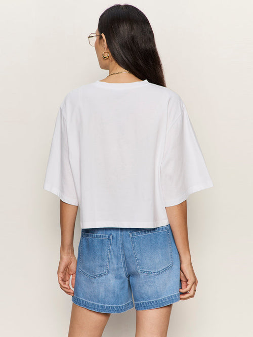 A woman with long dark hair and glasses stands with her back to the camera against a plain light background, wearing the Sanctuary Clothing "belle tee white" paired with high-waisted blue denim shorts.