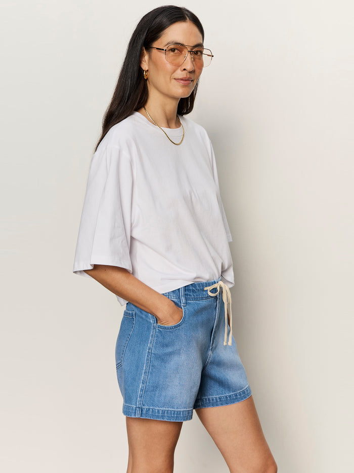 A woman with long dark hair, glasses, and gold jewelry wears the Sanctuary Clothing belle tee in white and denim drawstring shorts, standing with hands in pockets against a plain light background.