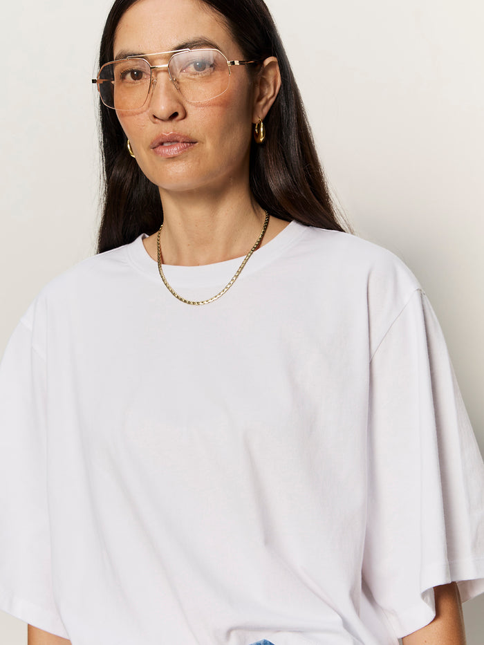 A woman with long dark hair wears Sanctuary Clothing's belle tee in white, paired with gold aviator glasses, gold hoop earrings, and a gold chain necklace, standing against a plain light background.