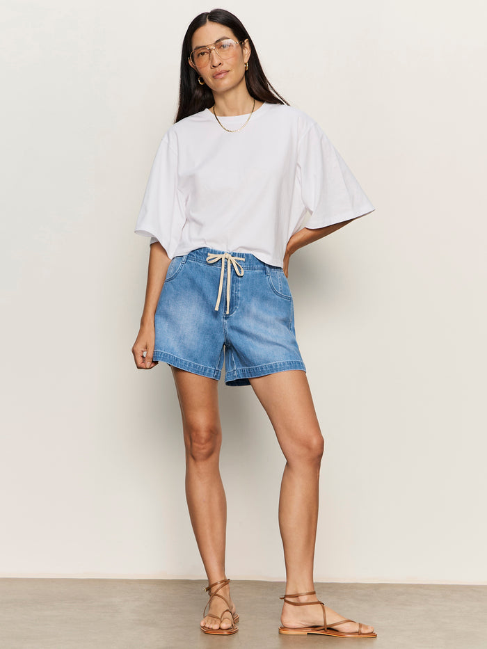 A woman with long dark hair stands against a plain background wearing the Sanctuary Clothing belle tee white, denim drawstring shorts, tan sandals, glasses, and a gold necklace. Her hands are in her pockets and she looks relaxed.