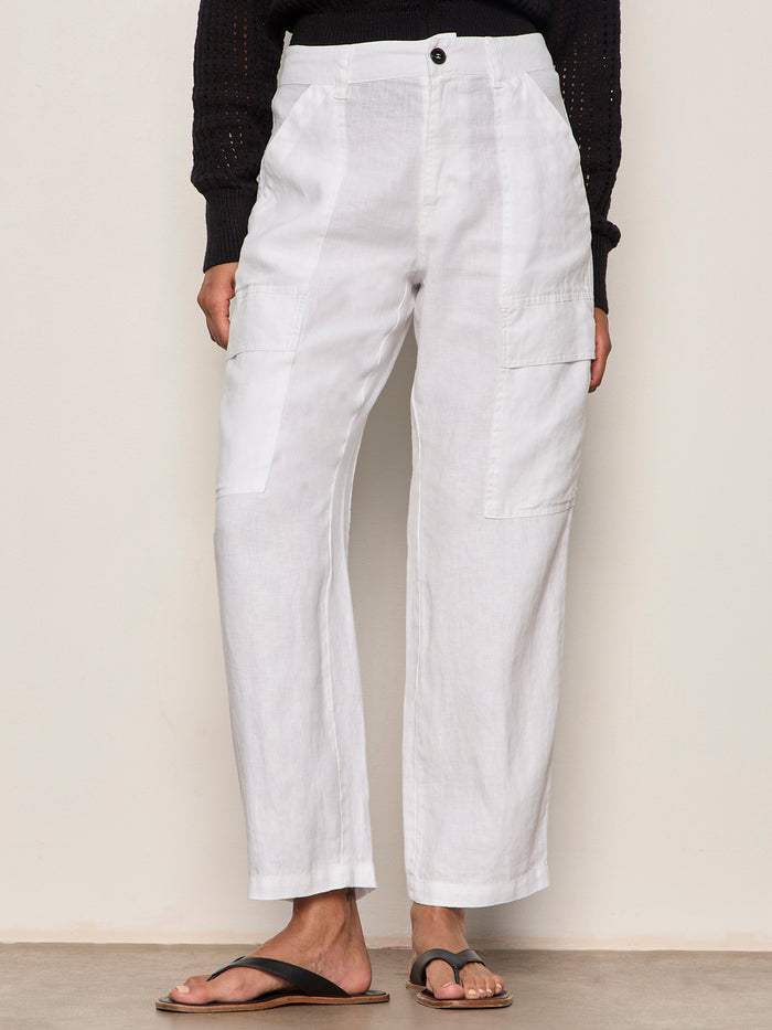 A person wears the Sanctuary Clothing boyfriend utility pant white 1—high-waisted linen pants with front and side pockets—paired with a black long-sleeve top and black sandals, standing against a plain light background.