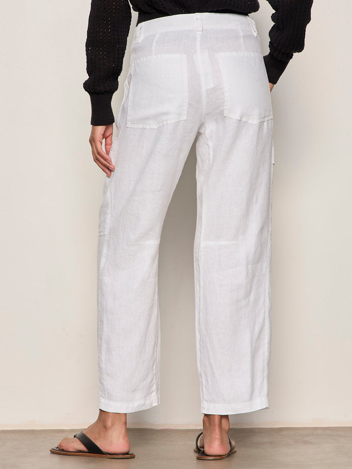 Wearing Sanctuary Clothing’s boyfriend utility pant white 1 with large back pockets, a black knit long-sleeve top, and black slide sandals, a person stands facing away from the camera against a light background.