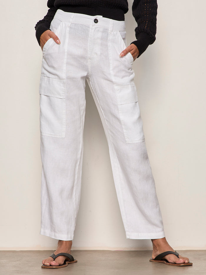 A person wears Sanctuary Clothing's boyfriend utility pant white 1, featuring large front pockets, paired with a black long-sleeve top and black sandals, standing against a light wall and floor with hands in their pockets.
