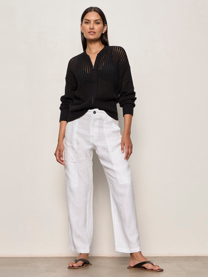 A person with straight dark hair and a neutral expression stands against a light wall, wearing loose-fitting Sanctuary Clothing boyfriend utility pant white 1, a black perforated long-sleeve sweater, and black sandals.