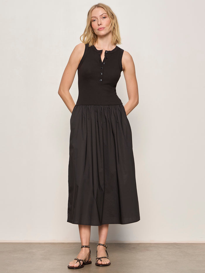 A woman with shoulder-length blonde hair stands by a light wall, wearing Sanctuary Clothing's breakaway mix media midi dress in black and black sandals. The sleeveless dress features a fitted top, button details, and a gathered skirt.