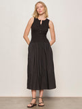 A woman with shoulder-length blonde hair stands by a light wall, wearing Sanctuary Clothing's breakaway mix media midi dress in black and black sandals. The sleeveless dress features a fitted top, button details, and a gathered skirt.