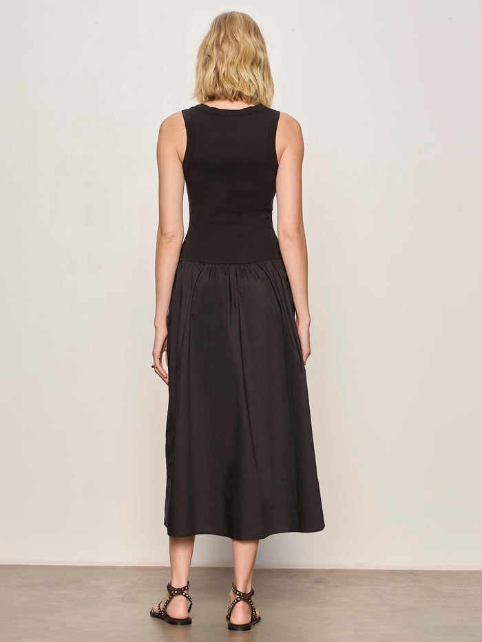 A woman with blonde hair wears the Sanctuary Clothing breakaway mix media midi dress in black, standing with her back to the camera against a plain light wall. She completes the look with flat, embellished sandals.