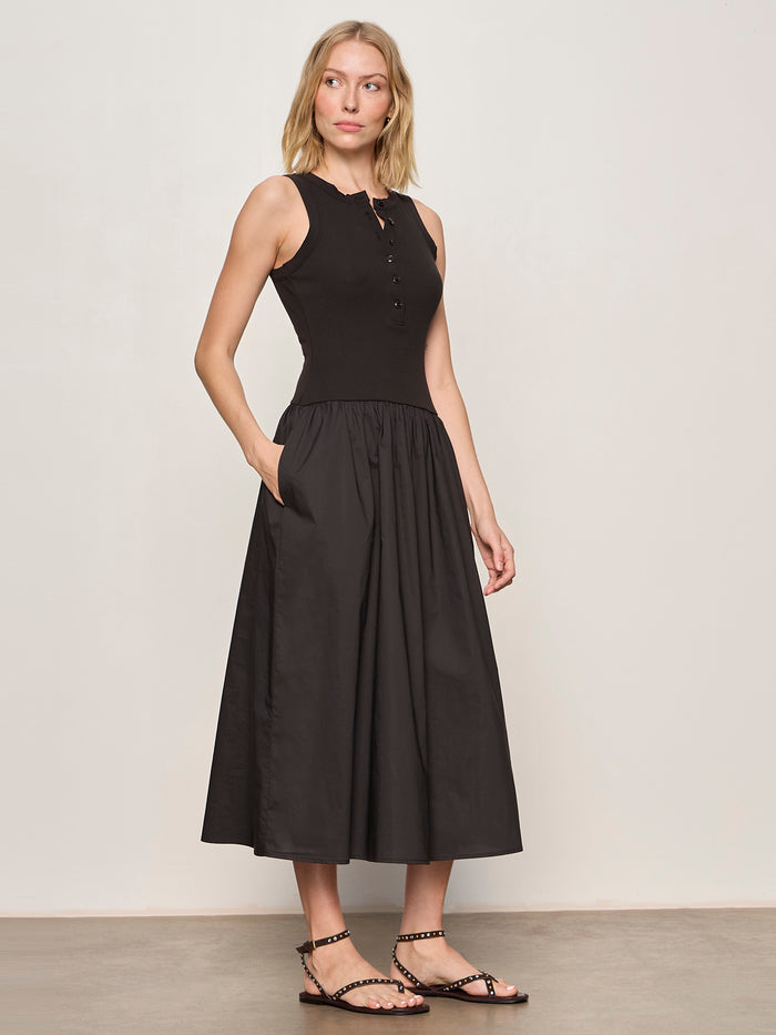 A woman with blonde hair stands against a plain background wearing the Sanctuary Clothing breakaway mix media midi dress in black, featuring a sleeveless fitted top and flared skirt. She pairs it with studded black sandals and has her hand in her pocket.