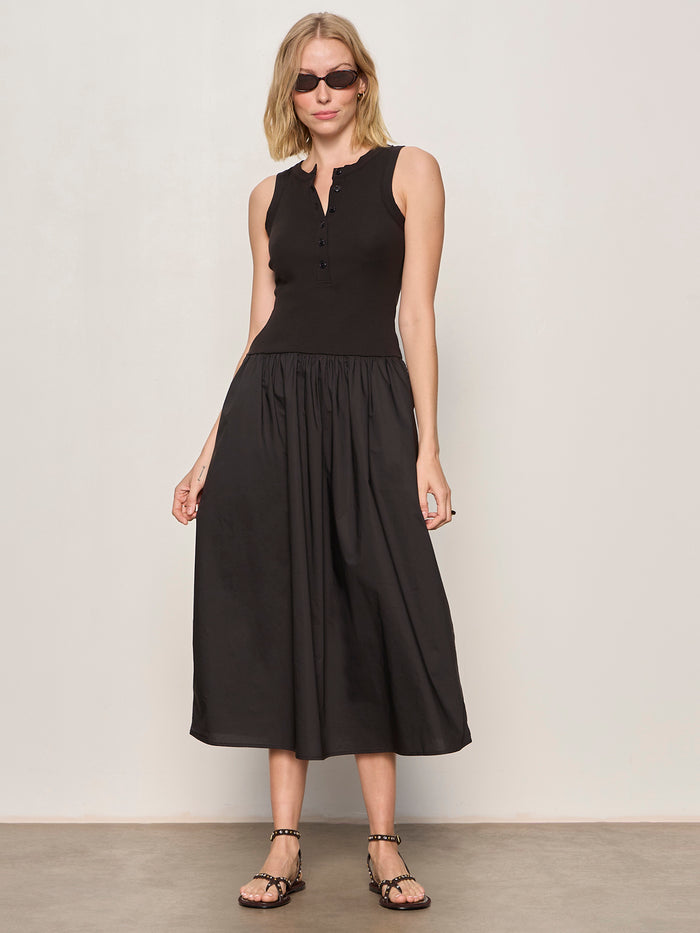 A woman stands against a plain light-colored wall, wearing Sanctuary Clothing's breakaway mix media midi dress in black with sunglasses and black sandals, her hands resting at her sides.