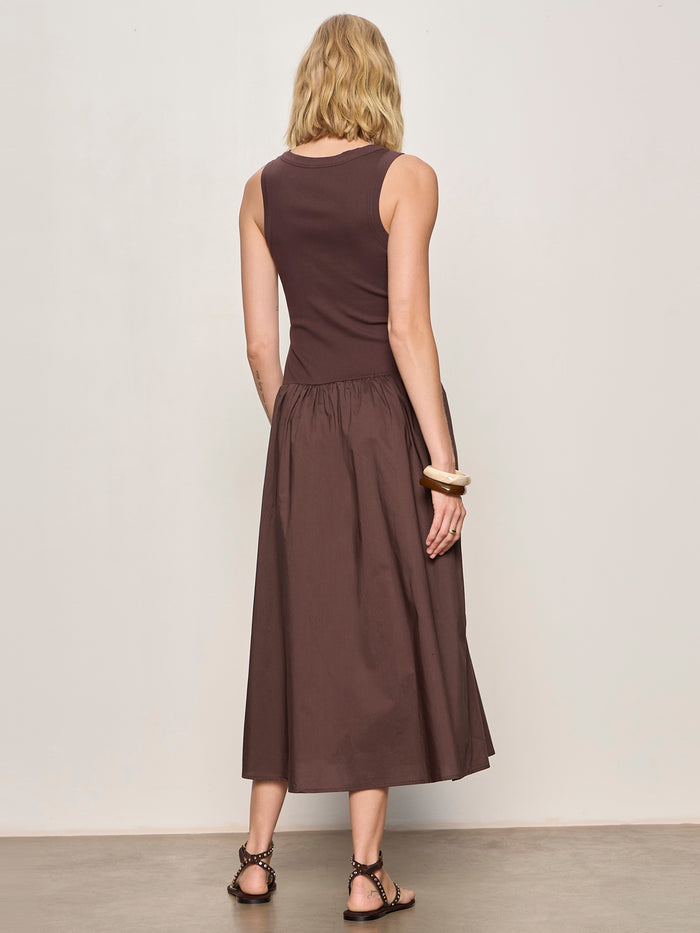 A woman with wavy blonde hair models the Sanctuary Clothing breakaway mix media midi dress in rich soil, facing away from the camera against a plain light beige background. She accessorizes with black sandals and large bracelets.