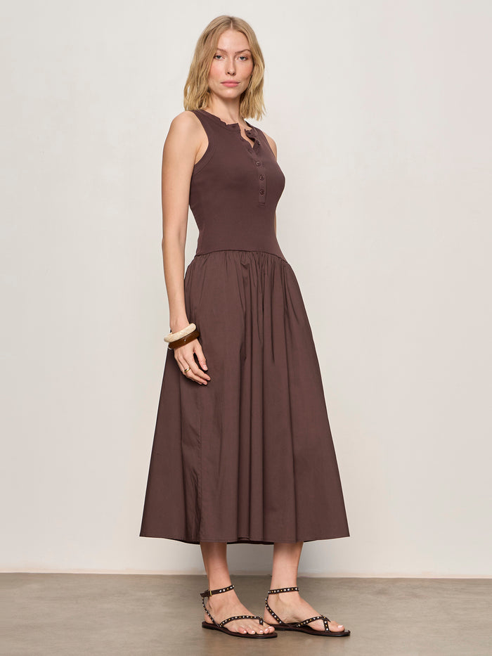 A woman with blonde hair and a neutral expression stands against a plain wall, wearing Sanctuary Clothing's breakaway mix media midi dress in rich soil, paired with black studded sandals and chunky bracelets.