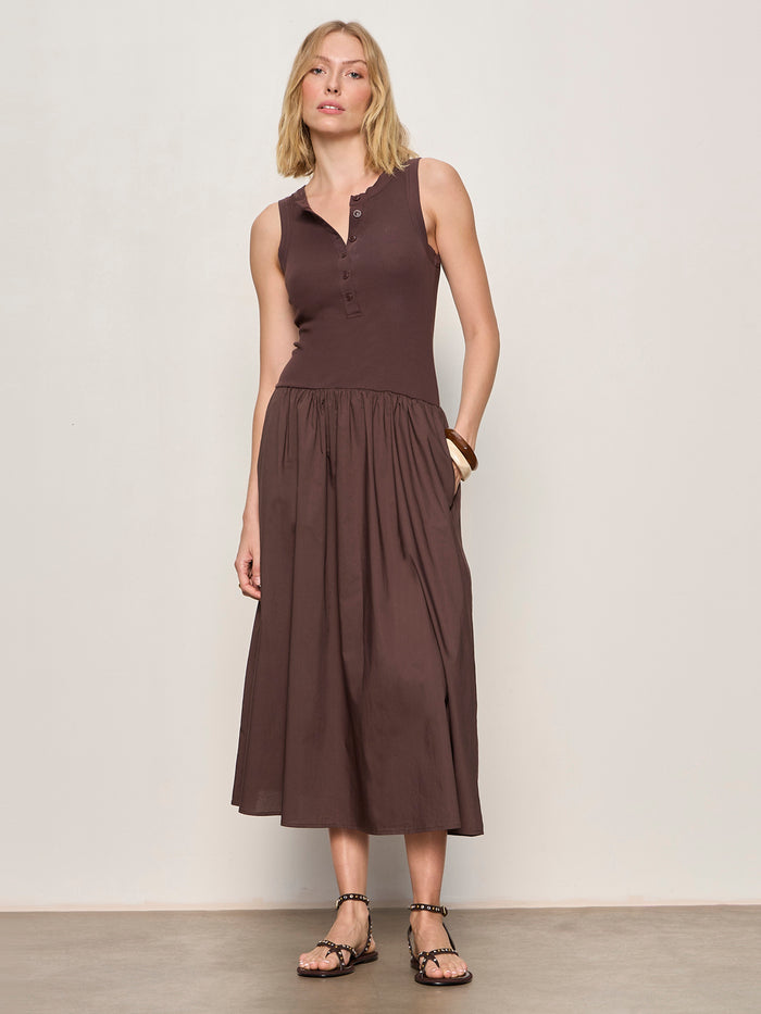 A woman with short blonde hair stands against a plain light wall, wearing Sanctuary Clothing's breakaway mix media midi dress in rich soil. She pairs the sleeveless, buttoned dress with flat sandals and a bracelet for a relaxed, casual look.