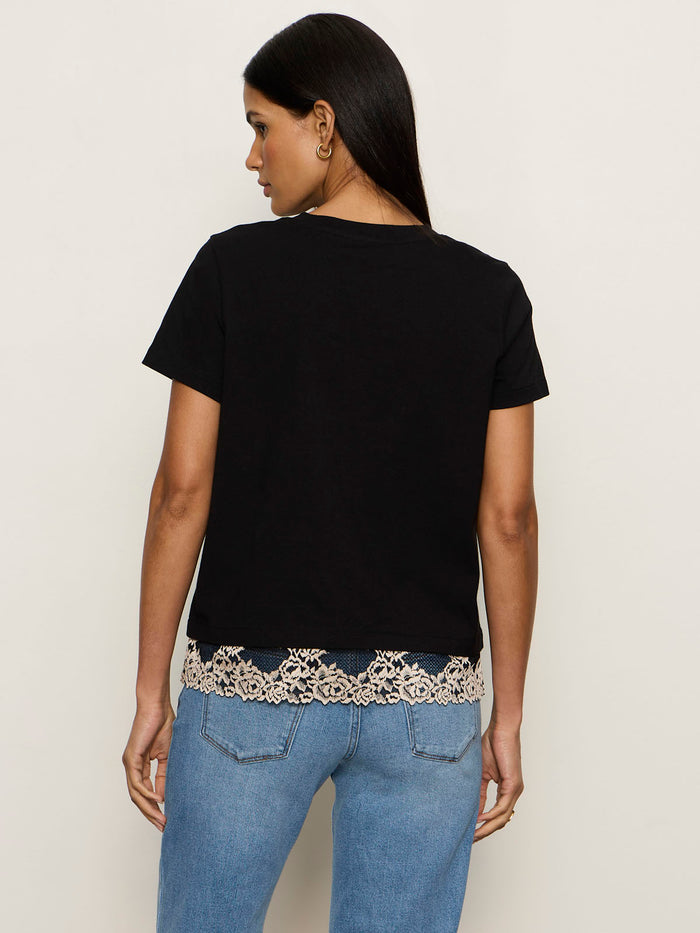 A woman stands with her back to the camera, wearing the cami remix tee black by Sanctuary Clothing, featuring beige lace trim at the hem and paired with light blue jeans against a plain light background.