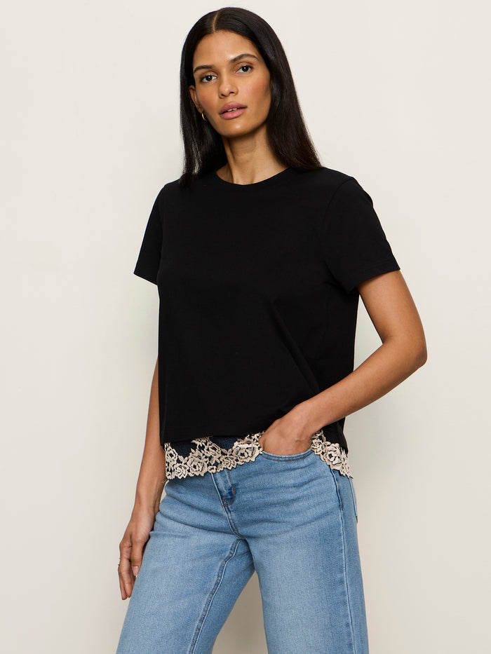 A woman with long dark hair wears the Sanctuary Clothing cami remix tee in black, featuring beige lace trim at the hem, paired with light blue jeans and standing against a plain light-colored background.