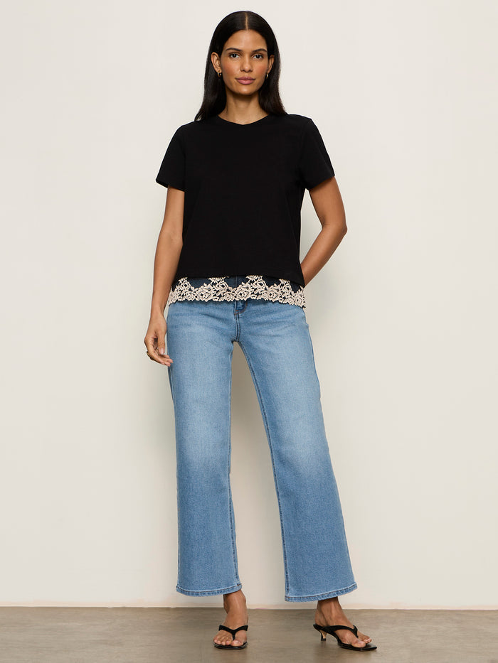 A woman with long dark hair and a neutral expression stands against a plain wall wearing the Sanctuary Clothing cami remix tee in black, paired with light blue wide-leg jeans and black heeled sandals. Her arms are relaxed by her sides.