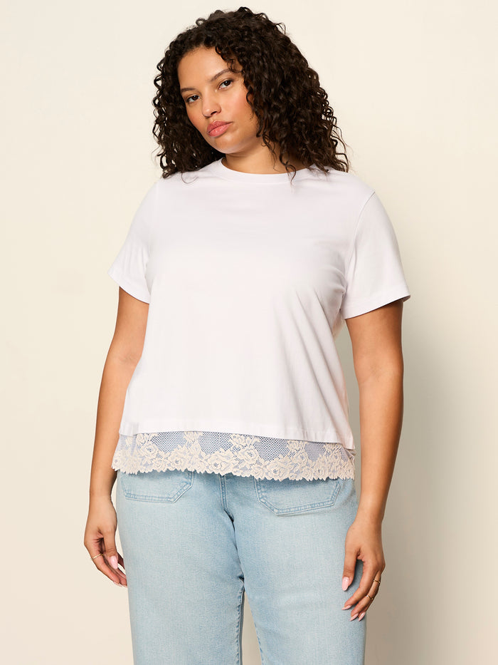 A woman wears the Sanctuary Clothing cami remix tee in white (extended sizing) and light blue jeans, standing against a beige background and looking at the camera with a neutral expression.