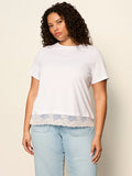 A woman wears the Sanctuary Clothing cami remix tee in white (extended sizing) and light blue jeans, standing against a beige background and looking at the camera with a neutral expression.