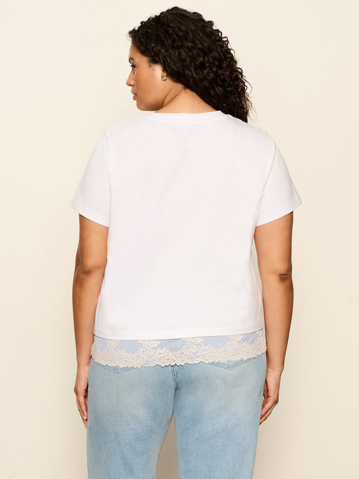 A woman with curly hair, seen from behind, wears the Sanctuary Clothing cami remix tee in white extended sizing and light blue jeans against a plain beige background.