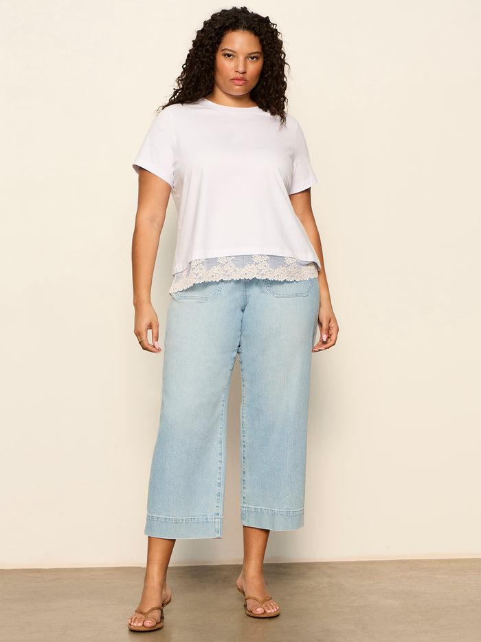 A person with curly hair models the Sanctuary Clothing cami remix tee in white (extended sizing), paired with light blue wide-leg jeans and tan sandals, standing against a beige wall on a concrete floor.