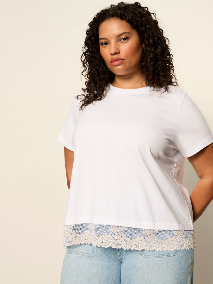 A woman with curly hair wears the Sanctuary Clothing cami remix tee white extended sizing and light blue jeans, standing against a neutral background.