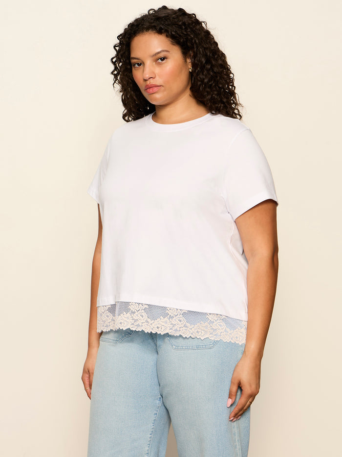A woman with curly hair wears Sanctuary Clothing's cami remix tee in white (extended sizing) and light blue jeans, standing against a plain beige background.