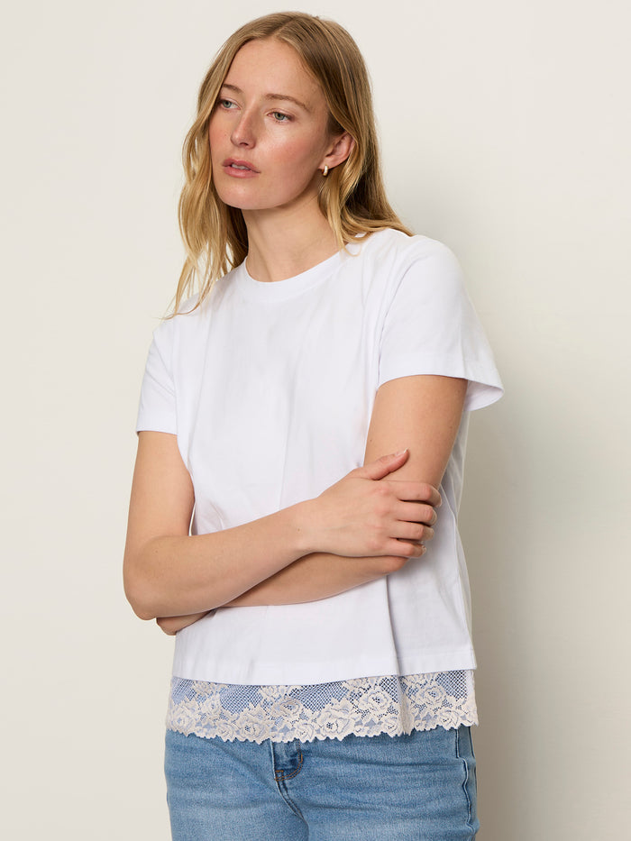 A woman with long blonde hair wears the Sanctuary Clothing cami remix tee in white, featuring lace trim at the hem, paired with blue jeans. She stands against a light background, looking to the side with arms loosely crossed.
