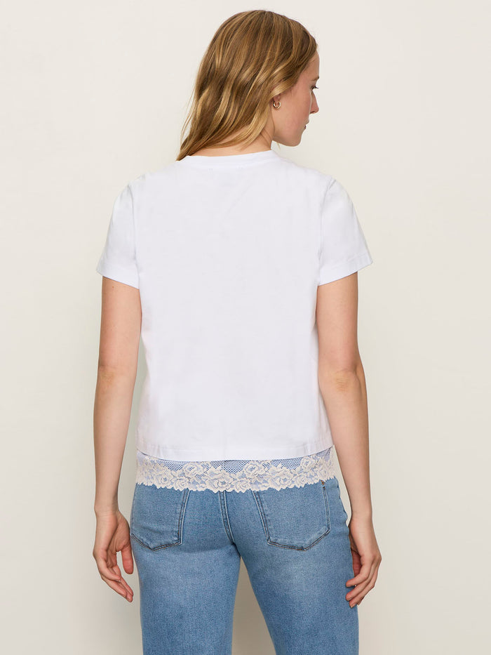 A woman with long blonde hair stands with her back to the camera in a Sanctuary Clothing cami remix tee white and light blue jeans, against a plain, light background.