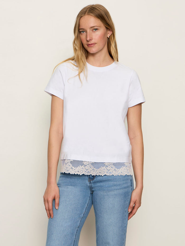 A woman with long blonde hair models the Sanctuary Clothing cami remix tee in white, featuring lace trim at the hem, paired with light blue jeans against a neutral background.