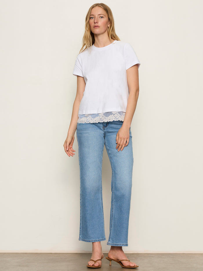 A woman stands against a plain background wearing the Sanctuary Clothing cami remix tee in white, paired with light blue straight-leg jeans and tan heeled sandals.