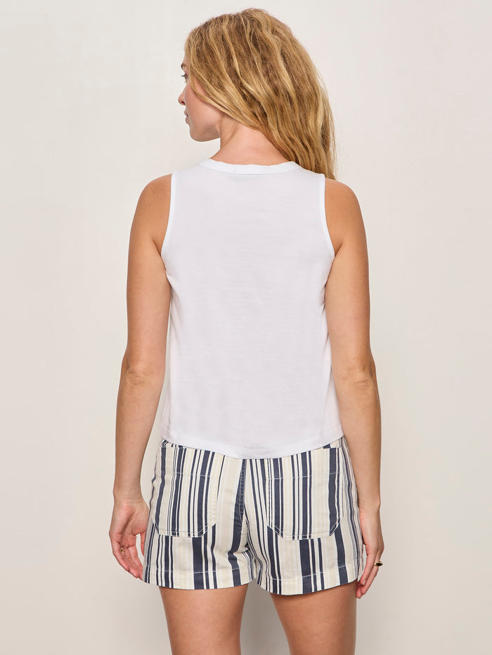 A woman with long, wavy blonde hair stands with her back to the camera in a Sanctuary Clothing campari embellished tank in white. She pairs it with beige shorts featuring vertical navy and white stripes against a plain, light backdrop.